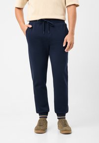 Námořnické modré tepláky vyrobené z měkké látky, s elastickým pasem se šňůrkou a žebrovanými manžetami. Stylizované s bočními kapsami.