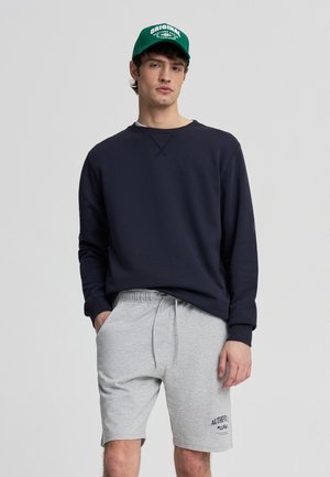 Giovane uomo che indossa un cappellino verde, una felpa blu navy e pantaloncini grigi chiaro con coulisse, con la scritta "Authentic" su una gamba, in piedi davanti a uno sfondo uniforme.