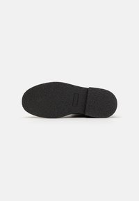 Semelle de chaussure en caoutchouc noir avec une surface texturée et une légère courbure au niveau du talon. Présente une étiquette rectangulaire près de l'arrière.