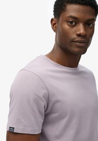 T-shirt en coton lilas clair avec des manches courtes, un col rond et une petite étiquette de marque cousue sur la manche gauche. Texture lisse.