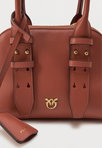 Τσάντα χρώματος σκουριάς από δέρμα με δύο λουριά, χρυσό φερμουάρ, χρυσό λογότυπο πουλιού και αποσπώμενη δερμάτινη ετικέτα που είναι αποκρουσμένη με το "PINKO" σε άσπρο φόντο.