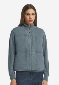 Helles blaues Sweatshirt mit Reißverschluss, Kabelstrickmuster auf der Vorderseite, hohem Kragen und langen Ärmeln, aus weichem Strickstoff gefertigt.