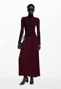 Bordeauxrode geribbelde coltrui lange jurk met een geplooide rok, gecombineerd met een zwarte zonnebril en een marineblauwe handtas. Strakke, aangesloten pasvorm.