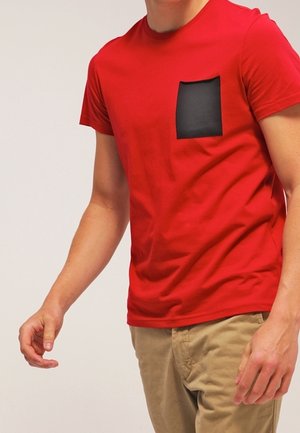 Man in een rood T-shirt met korte mouwen en een zwarte borstzak, met beige broek, staand tegen een effen achtergrond.