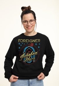 Felpa nera con un design grafico di un jukebox, con testo multicolore che legge "Foreigner Jukebox Hero" e decorazioni a forma di stella.