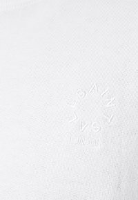 Camiseta de algodón blanca de canalé con un pequeño diseño de sol en relieve en la parte izquierda del pecho, con sutil textura y detalles de costura.