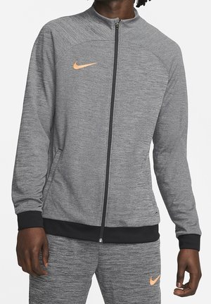 Mann trägt eine graue Nike-Reißverschlussjacke und passende Hose mit orangefarbenen Nike-Swoosh-Logos, steht vor einem einfarbigen Hintergrund.