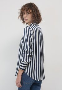 Camicetta a righe nei colori blu navy e bianco, tessuto semi-trasparente, maniche lunghe e orlo posteriore allungato. Indossata con jeans blu.