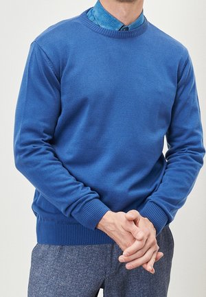 Pull bleu à col rond en tissu tricoté doux. Bords-côtes aux poignets et à l'ourlet, superposé à une chemise à col bleu clair.