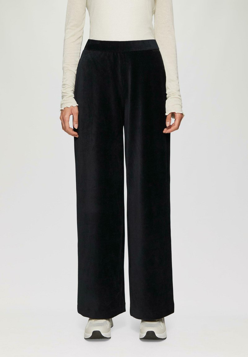 Zwarte fluwelen broek met brede pijpen, een gladde textuur en een elastische tailleband. Gecombineerd met een licht, langmouwtop en sneakers voor een casual uitstraling.