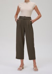 Haut beige côtelé, pantalon taille haute vert olive sur mesure avec ceinture et plis. Le mannequin porte des talons blancs à brides, sur un fond gris clair.
