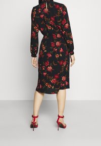 Robe midi fleurie en tissu noir, ornée de motifs colorés de roses, manches longues, taille cintrée et fente au dos. Associée à des escarpins rouges.