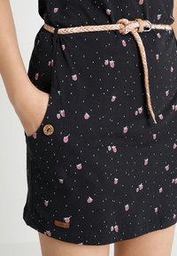 Personne portant une jupe noire avec un petit imprimé de fruits rouges, une poche latérale boutonnée et une ceinture tressée beige nouée à la taille.