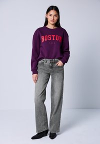 Lila Sweatshirt mit rotem Aufdruck "BOSTON 1980", kombiniert mit hellgrauen geraden Jeans und schwarzen, hohen Stiefeletten.
