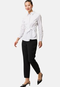 Vit skjorta med knytning i midjan, kombinerad med smala svarta byxor och svarta slingback-skor. Slät tygkvalitet, strukturerad design.