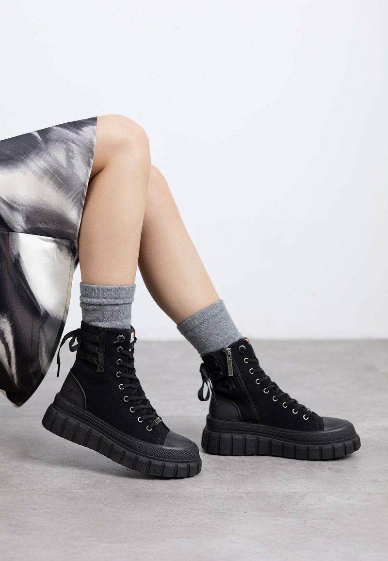 Zapatillas altas negras de goma y tela, con una suela gruesa y texturizada, diseño de cordones y un detalle de cremallera lateral. Calcetines grises visibles.