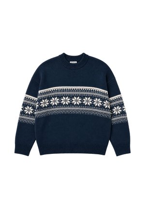 Mand iført en grå strikket hue og en marineblå sweater med hvide snefnugsmønstre, stående mod en ensartet lys baggrund.