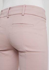 Close-up van lichtroze op maat gemaakte broeken met riemlussen, achterzakken met bies, gedragen met een wit topje.