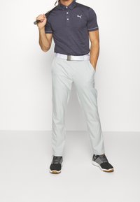 Pánská polokošile ve tmavě modré barvě, s malým logem, kombinovaná se světle šedými kalhotami a černými sportovními botami. Hladká tkanina, strukturovaný střih.