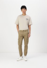Beige T-shirt met geprinte accenten, kaki cargobroek met zijzakken en witte sneakers. Slim fit, lichte stof, casual ontwerp.