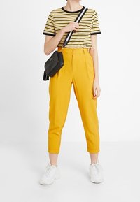 Gula, höga byxor med en smal passform, kombinerade med en svart och gul randig t-shirt. En liten svart axelremsväska och vita sneakers.