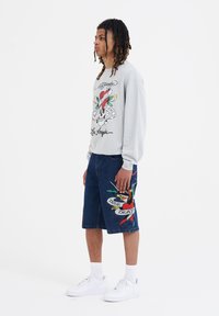 Sudadera gris con gráfico y texto colorido, combinada con pantalones cortos de mezclilla azules que presentan un vívido diseño bordado. Zapatillas blancas.