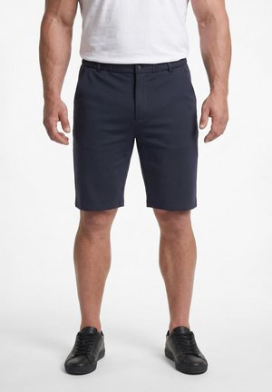 Mand iført marineblå skræddersyede shorts, hvid t-shirt og sorte sneakers stående mod en helt hvid baggrund.