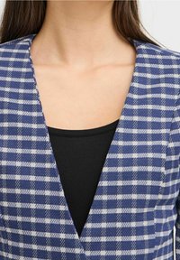 Femme portant un blazer à carreaux bleu et blanc sur un haut noir, montrant le décolleté et le haut de la poitrine avec des cheveux bruns raides.