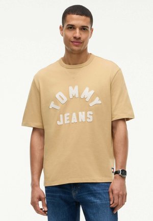 Mand iført beige Tommy Jeans t-shirt og blå jeans, stående med et neutralt udtryk mod en ensfarvet baggrund.