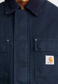 Marineblaue Jacke mit Cordkragen, zwei Vordertaschen und Carhartt-Logo-Tag. Hergestellt aus strapazierfähigem Stoff mit sichtbaren Nähten.