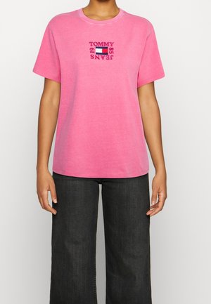 Kobieta w różowej koszulce z krótkim rękawem z napisem „Tommy Jeans 1985” i logo flagi na piersi, połączona z czarnymi szerokimi spodniami.