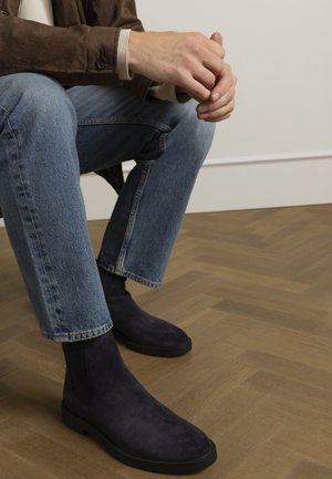 Marineblauwe suède chelsea boots met een gladde textuur en een minimalistisch ontwerp, gecombineerd met lichtblauwe jeans en een bruine jasje, geplaatst op een houten vloer.