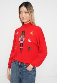Czerwony sweter z dzianiny z motywem dziadka do orzechów, zdobiony cekinami i dodatkami, okrągłym dekoltem oraz luźnym krojem. Akcenty to płatki śniegu i gwiazdy.