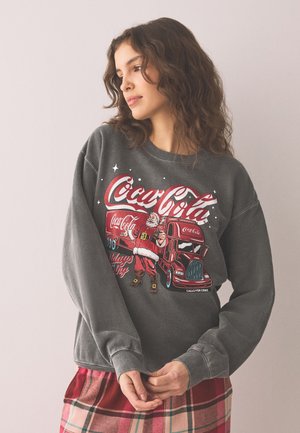 Jonge vrouw draagt een grijze Coca-Cola sweater met de kerstman en een rode vrachtwagen afbeelding, gecombineerd met een rood en roze geruite rok.