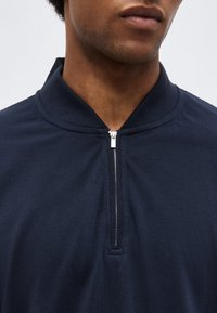 Gros plan sur une personne portant une chemise à col zippé bleu marine, avec la fermeture éclair partiellement fermée au niveau du cou.