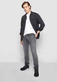 Tânăr stând cu mâna în buzunar, îmbrăcat cu o jachetă din denim negru, un tricou alb, blugi gri și adidași negri cu glezna, pe un fundal gri.