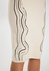 Fustă tip creion tricotată în nuanță crem, decorată cu linii ondulate de paiete negre, argintii și aurii, creând o textură și un design intricate.