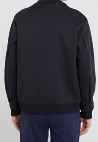 Sudadera negra con cuello y puños acanalados, corte recto y textura suave. Detalle de costura visible en el dobladillo.
