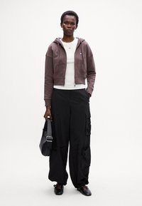 Brun cropped hette genser over en hvit tettsittende topp, kombinert med svarte cargo bukser. Tilbehøret inkluderer en svart skulderveske og svarte sko.