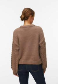 Femme aux cheveux bruns coiffés en chignon, portant un pull en maille torsadée marron et un jean bleu foncé, vue de dos sur un fond blanc.