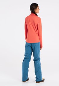 Koraalrode top met lange mouwen en hoge kraag, gecombineerd met blauwe, losse broek. De outfit is gemaakt van zachte, flexibele materialen voor buitenactiviteiten.