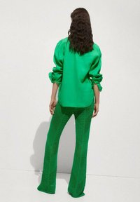 Bluză de mătase verde cu mâneci bufante, asociată cu pantaloni evazați din țesătură de plasă, ambele având o nuanță verde uniformă și texturi unice.