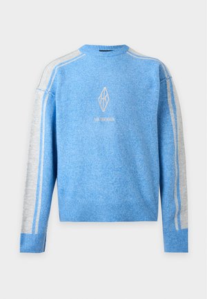 Junger Mann steht, trägt einen blau-grauen Langarmpullover mit braunen Cargohosen und weißen Turnschuhen vor weißem Hintergrund.