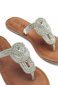 Sandalias de dedo con cuentas plateadas y diseño anudado, con una plantilla de ante marrón y suela plana, que ofrecen un ajuste cómodo.