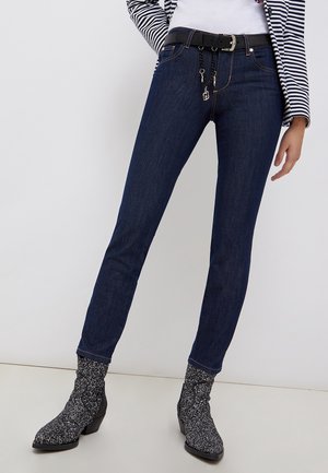 Mujer con vaqueros ajustados azul oscuro, cinturón negro con charms metálicos, blazer a rayas y botines moteados en blanco y negro.