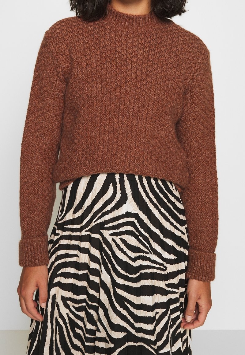 Femme portant un pull marron texturé et une jupe plissée à imprimé zèbre noir et blanc, debout devant un fond uni.