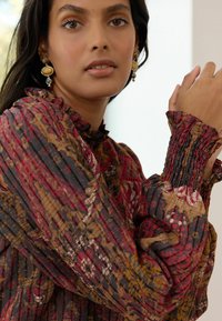 Multicolored blouse featuring intricate patterns in red, brown, and yellow. Ruffled collar, smocked cuffs, and lightweight pleated texture.