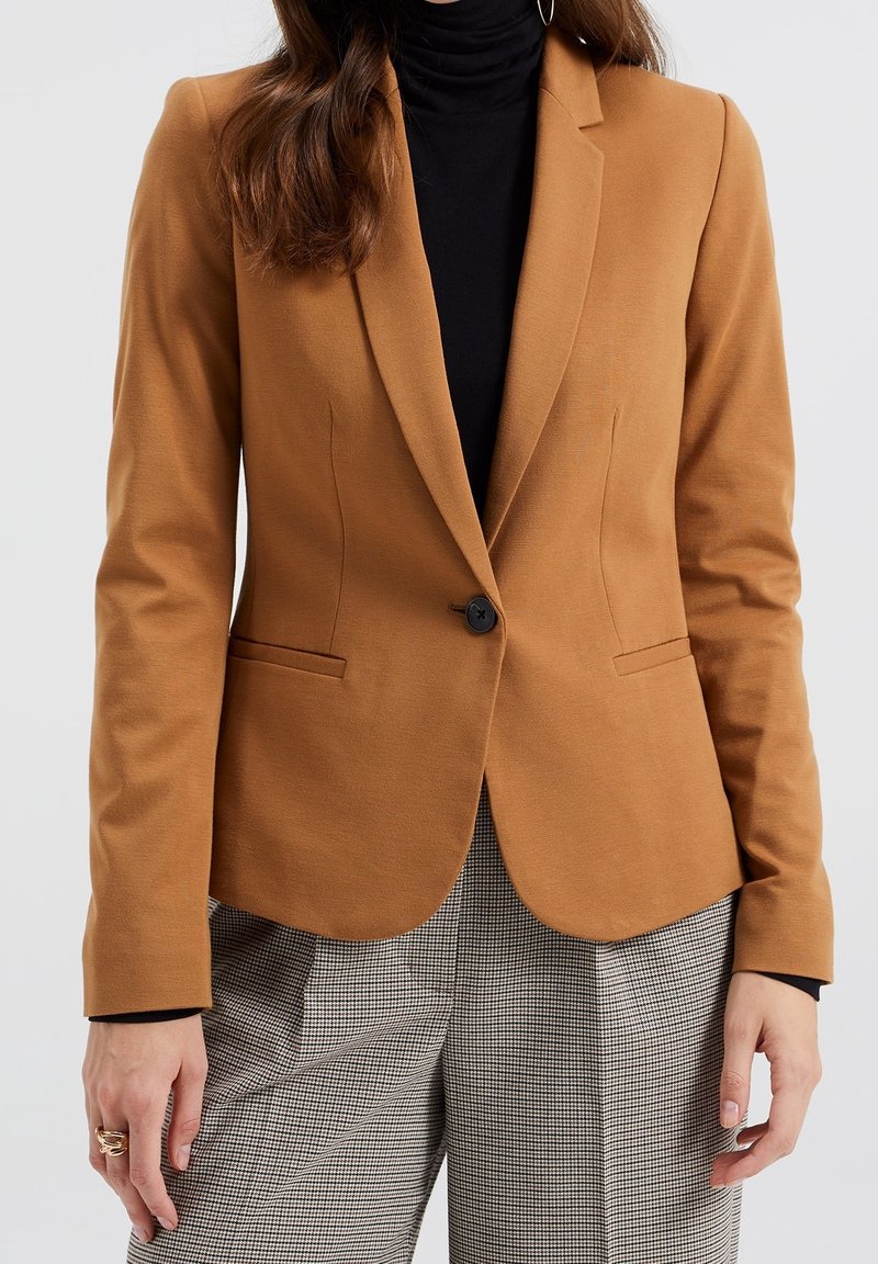 Personne portant un blazer couleur camel, un col roulé noir et un pantalon gris à carreaux, montrée des épaules aux cuisses devant un fond uni.