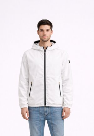 Uomo in piedi di fronte, indossa una giacca bianca con cappuccio e cerniera nera e jeans azzurri chiari su uno sfondo bianco semplice.