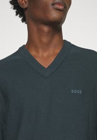 Homme portant un pull à col en V vert foncé avec le logo "BOSS" brodé sur la poitrine gauche, sur un fond uni.